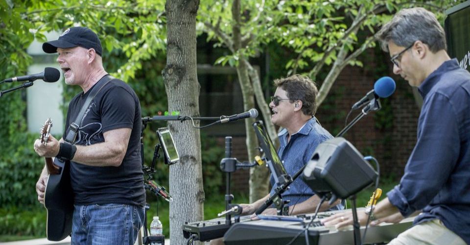 Band playing on stage outdoors with various instruments singing into microphone next to trees at SKYBOKX 109 Sports Bar & Grill in Natick, MA