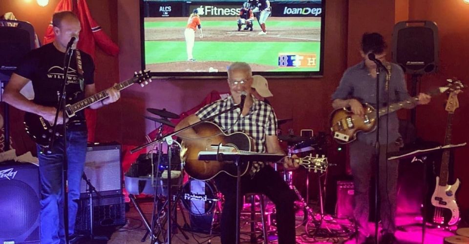 Band on stage with various instruments playing and singing into microphone in front of television playing sports near speakers at SKYBOKX 109 Sports Bar & Grill in Natick, MA